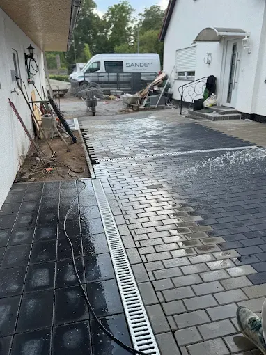 Dieselbe Einfahrt nach der Pflasterung: durchgehend verlegte Fläche mit dunkler Randeinfassung und einer Entwässerungsrinne entlang des Hauses.