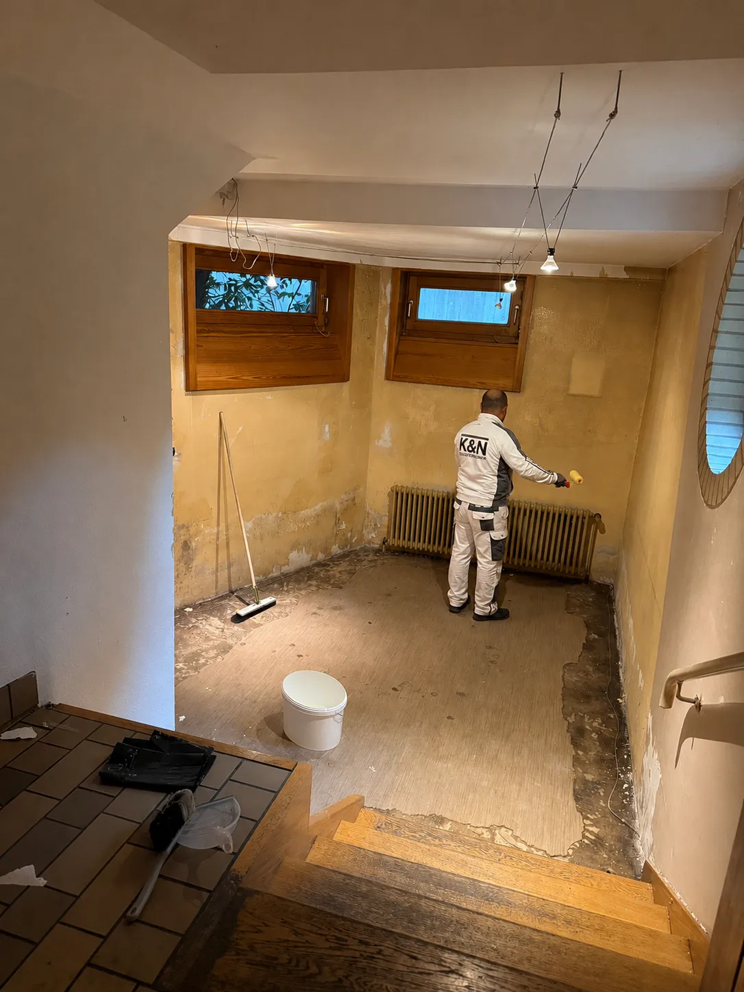 Kellerraum vor der Sanierung: Tapete entfernt, Wände fleckig und abgelaugt, Estrich freigelegt, Mitarbeiter von K&N beginnt mit den Malerarbeiten.